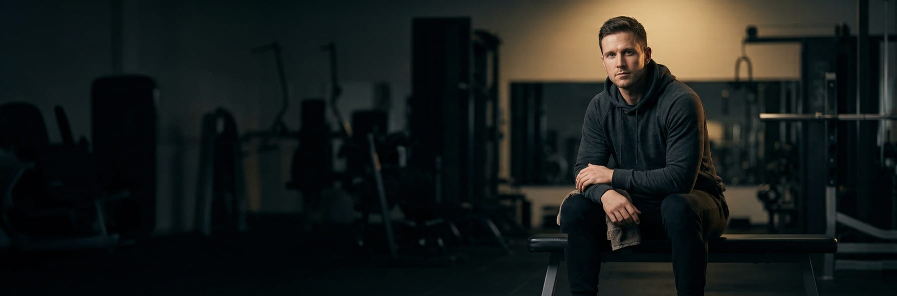 Lone athlete sitting on a gym bench in an empty dark gym — hands clasped, focused before lifting